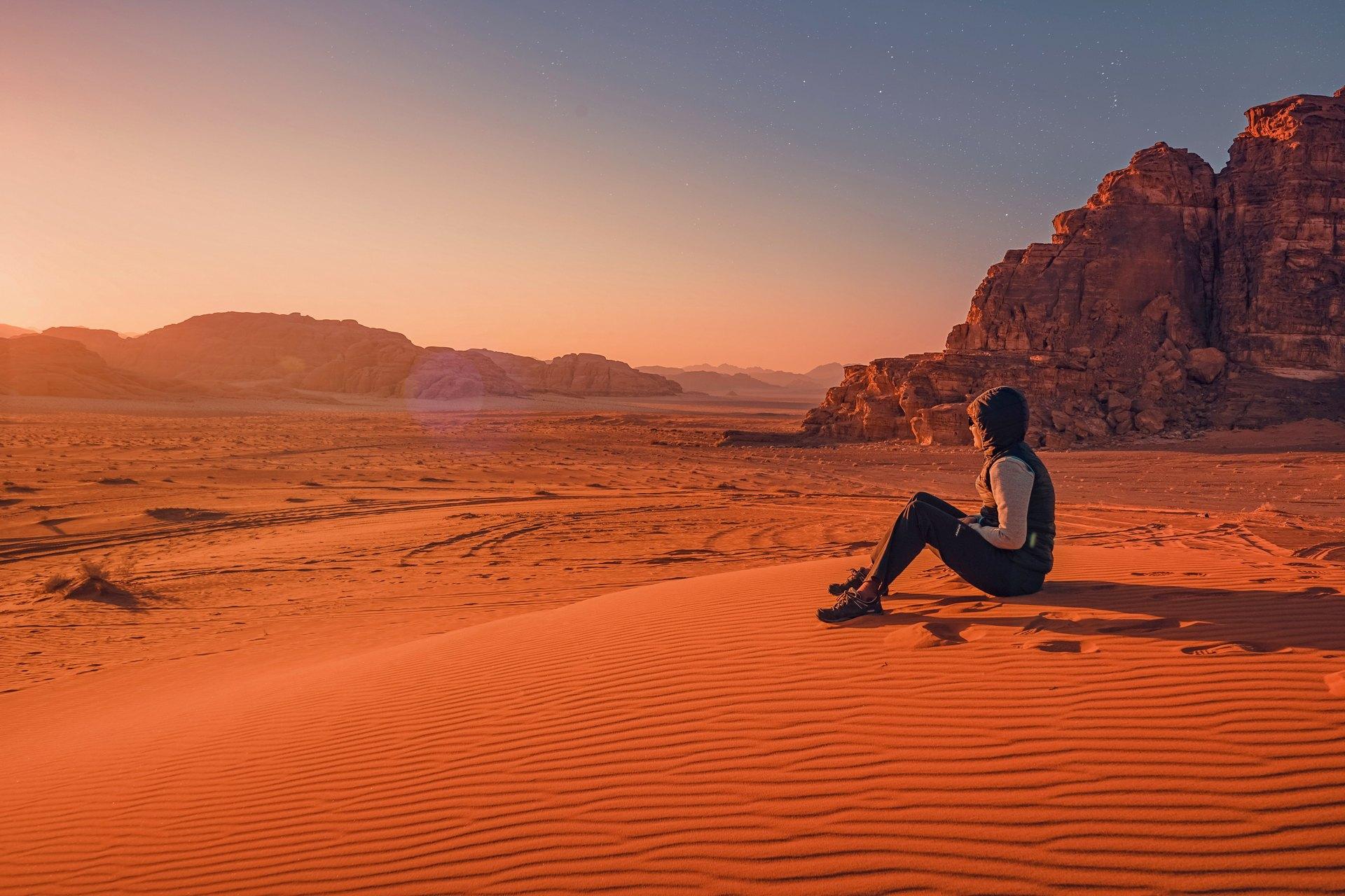 A man sitting on a sand dune in Wadi Rum, gazing at the vast desert landscape; A traveler resting on golden sands with dramatic sandstone mountains in the background; A lone explorer enjoying the serene beauty of Wadi Rum's desert at sunset; A man seated on a rippled sand dune, admiring the endless horizon of Wadi Rum; A peaceful moment in Wadi Rum with a traveler soaking in the breathtaking desert scenery; A visitor sitting atop a dune, overlooking the Mars-like terrain of Wadi Rum; A silhouette of a man on a sand dune against the warm hues of the setting sun in Wadi Rum; A contemplative traveler taking in the majestic rock formations and open desert of Wadi Rum; A man enjoying solitude on a sand dune with sweeping views of Wadi Rum’s untouched wilderness; A desert adventurer pausing on the dunes, surrounded by the raw beauty of Wadi Rum.